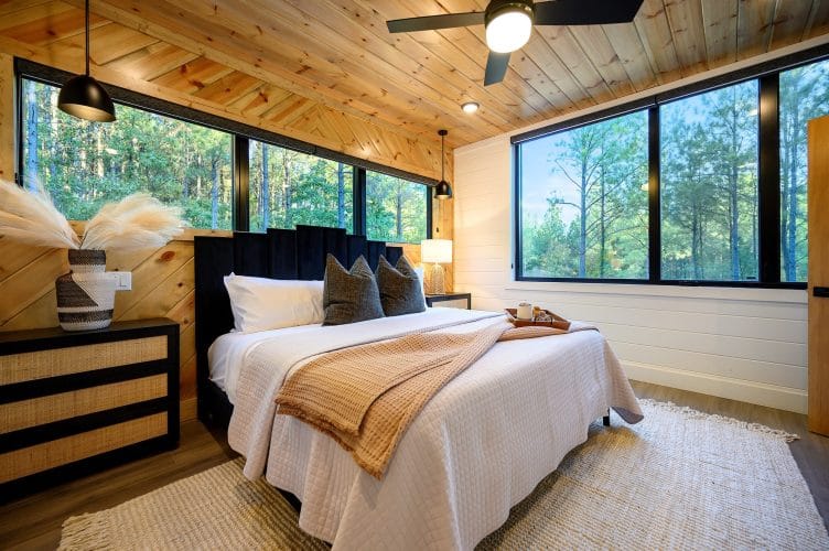 King bed with a black upholstered headboard under a pine plank ceiling, windows along the wall