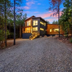 Front of the dark two-story cabin with cedar trim at dusk, a gravel drive and tall pines