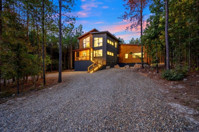 Front of the dark two-story cabin with cedar trim at dusk, a gravel drive and tall pines