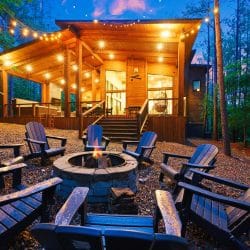 Fire pit ringed with dark Adirondack chairs, with the lit cabin above