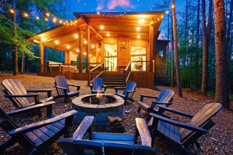 Fire pit ringed with dark Adirondack chairs, with the lit cabin above
