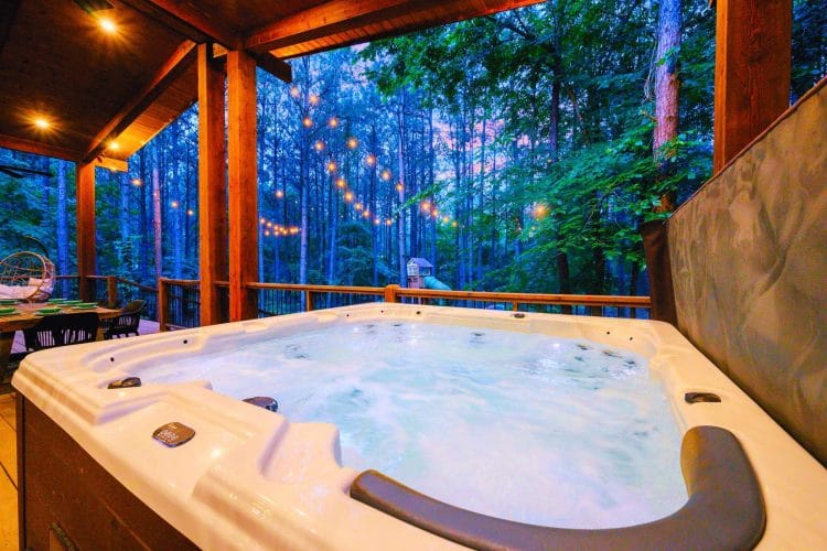 Hot tub on a covered deck among tall pines, with string lights beyond the rail