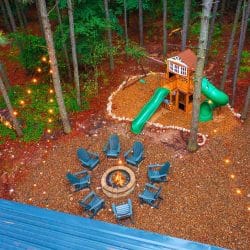 Aerial of the playset with a green slide, a ring of Adirondack chairs around a fire pit and string lights over red mulch