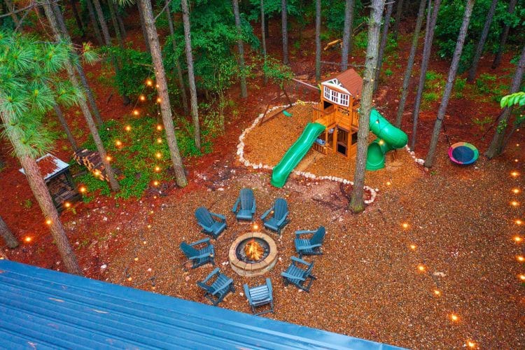 Aerial of the playset with a green slide, a ring of Adirondack chairs around a fire pit and string lights over red mulch