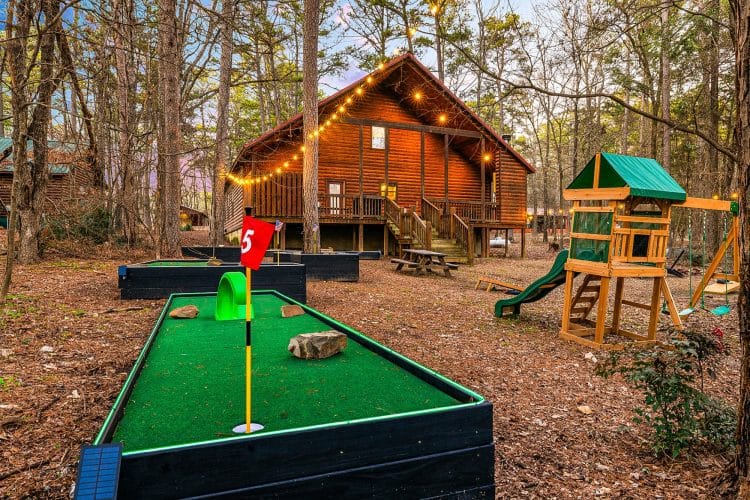 Mini golf hole with a red flag, the playset and the cabin behind
