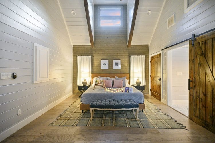 King bed under a wood accent wall with a tufted bench and plaid rug, the barn door open to the bath