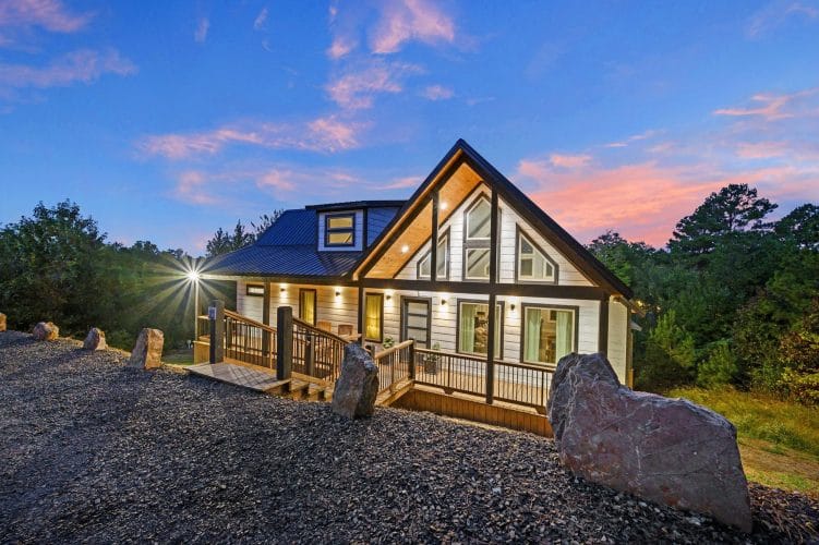 Front of the cabin with a tall glass gable, a deck and large boulders at dusk
