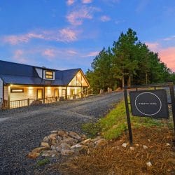 Cabin with a dark roof and glass gable above a gravel drive, with a black cabin-name sign at the roadside