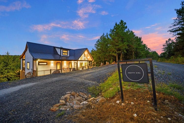 Cabin with a dark roof and glass gable above a gravel drive, with a black cabin-name sign at the roadside