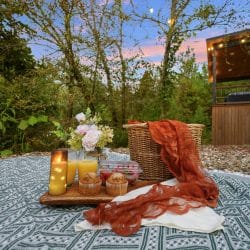 Picnic blanket with a basket and drinks on the gravel near the fire pit