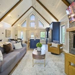 Open living room under a vaulted pine ceiling with exposed beams, a gas fireplace with a TV above it and a gray sofa
