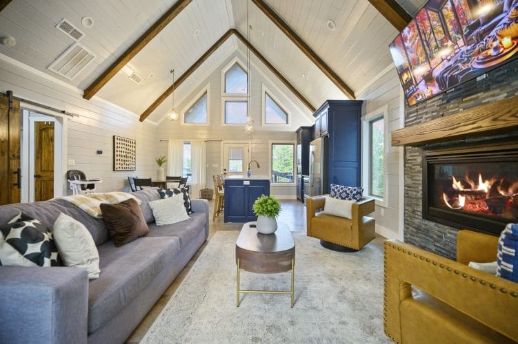 Open living room under a vaulted pine ceiling with exposed beams, a gas fireplace with a TV above it and a gray sofa