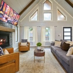 Living room with a gas fireplace and TV, a gray sofa, a tan leather chair and triangle windows