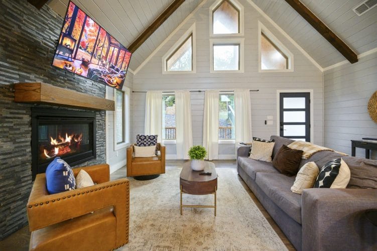 Living room with a gas fireplace and TV, a gray sofa, a tan leather chair and triangle windows