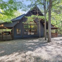 Dark blue-gray cabin with a covered porch and a gravel drive, seen through trees