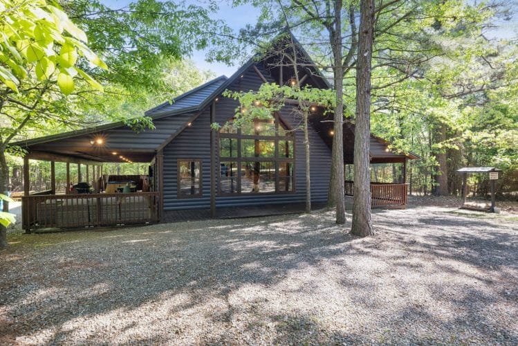 Dark blue-gray cabin with a covered porch and a gravel drive, seen through trees