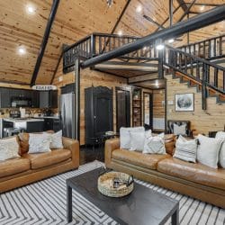 Living room with two tan leather sofas, a black coffee table and a striped rug, with the staircase and open kitchen behind