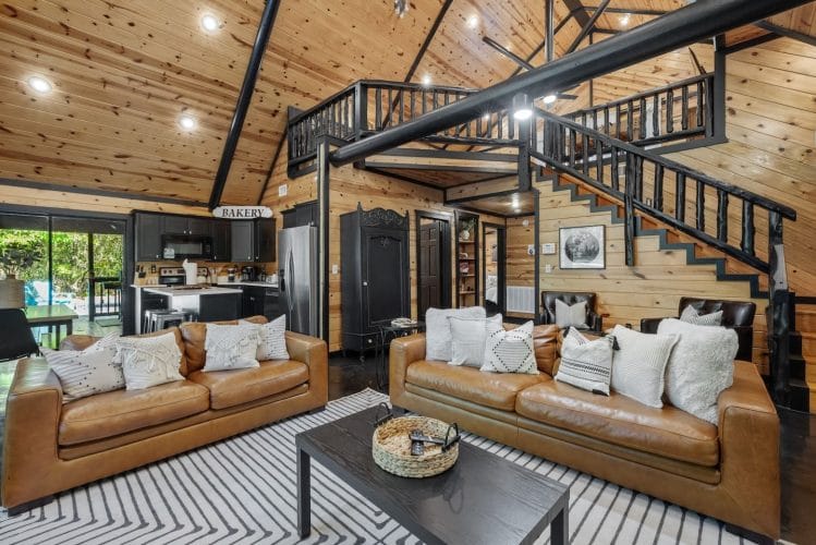 Living room with two tan leather sofas, a black coffee table and a striped rug, with the staircase and open kitchen behind