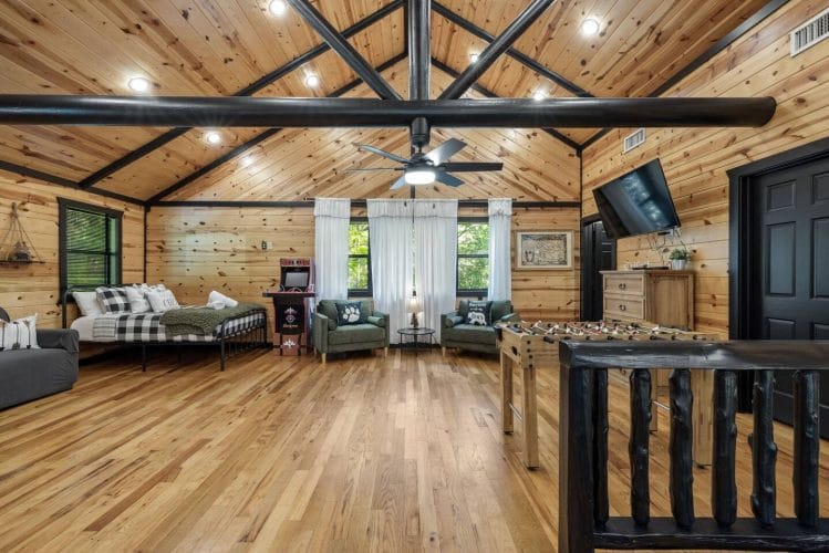 Open loft with a bed, a gray sofa, green armchairs, an arcade basketball game and a foosball table under a vaulted ceiling
