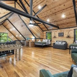 Loft with two beds in plaid bedding, a gray sofa, green armchairs and a desk, with a foosball table at the railing