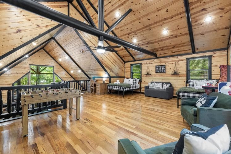 Loft with two beds in plaid bedding, a gray sofa, green armchairs and a desk, with a foosball table at the railing