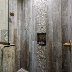 Shower niche with bottles next to a gray wood wall and river-stone tile
