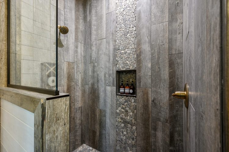 Shower niche with bottles next to a gray wood wall and river-stone tile