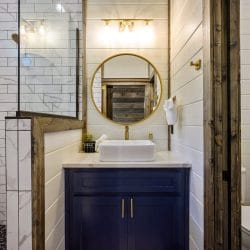Round gold mirror reflecting a reclaimed wood wall above a vessel sink