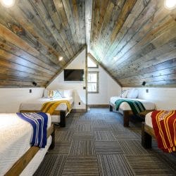 Upstairs bed with a yellow throw, a wall TV and a window under the vaulted ceiling