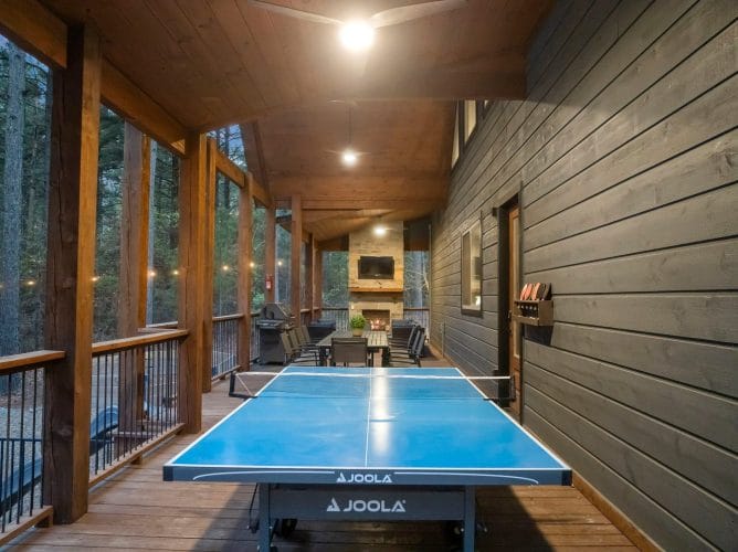 Covered porch at dusk with a dining table, a stone fireplace with TV and a grill