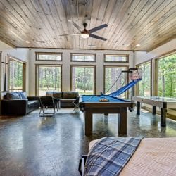 Blue-felt pool table by a window with a black leather couch and pine trees outside