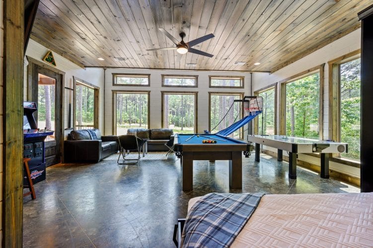Blue-felt pool table by a window with a black leather couch and pine trees outside