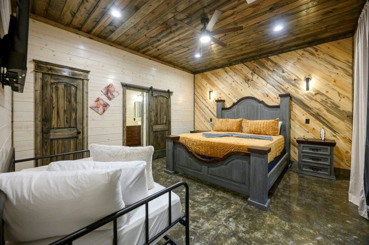 Foot of the bed with gold bedspread beside a wood door and the bathroom vanity
