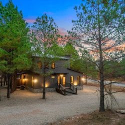 Dark-sided cabin at dusk with lit windows and pine trees around it
