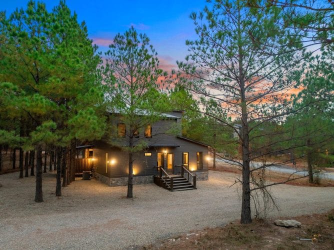 Dark-sided cabin at dusk with lit windows and pine trees around it