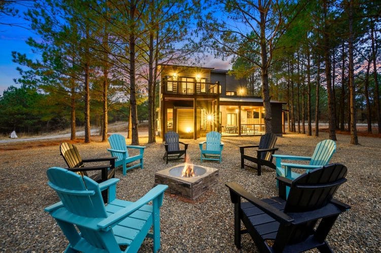 Fire pit glowing at night with teal and black Adirondack chairs and the lit patio behind