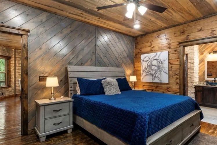 King bed with a navy quilt and gray wood headboard, nightstands with lamps, open door to the bathroom on the right