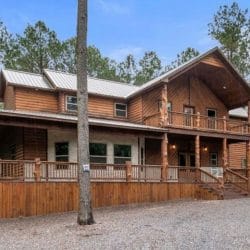 Two-story wood lodge with a wraparound front porch and a gravel drive among tall pines