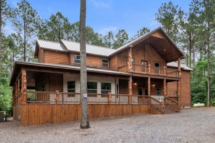 Two-story wood lodge with a wraparound front porch and a gravel drive among tall pines