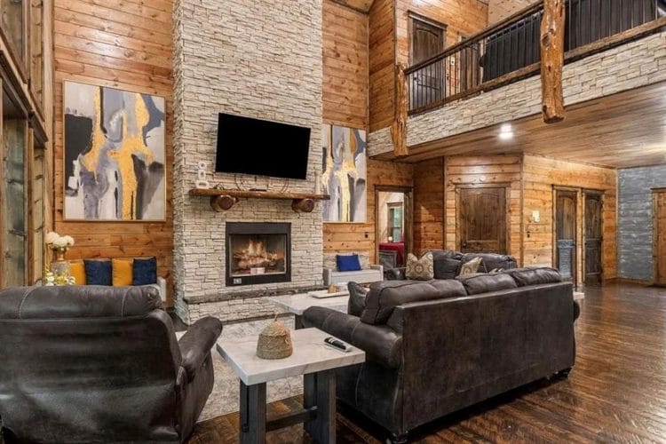 Living room with dark leather sofas and a stone fireplace with a TV above it, loft railing overhead