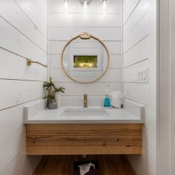 Small bathroom with a floating wood vanity and a round gold mirror, loft railing visible through the doorway