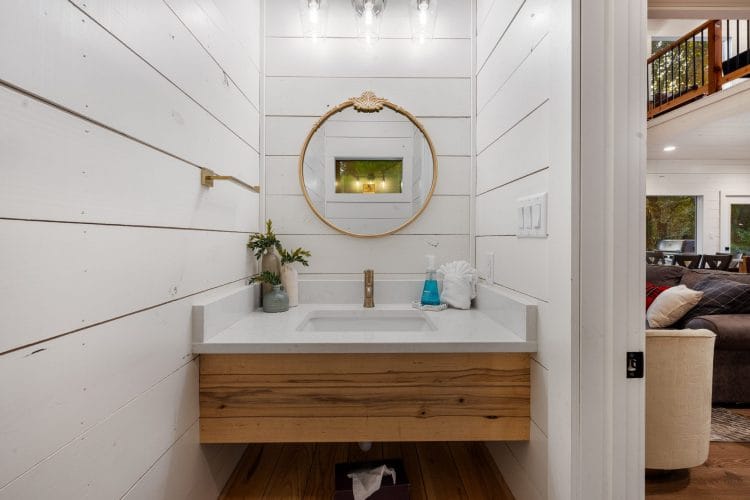 Small bathroom with a floating wood vanity and a round gold mirror, loft railing visible through the doorway