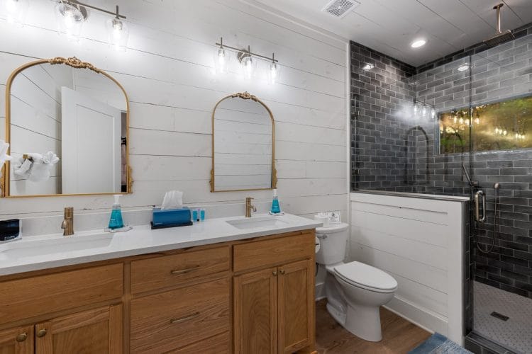 Double vanity with two arched gold mirrors and a glass walk-in shower with dark tile