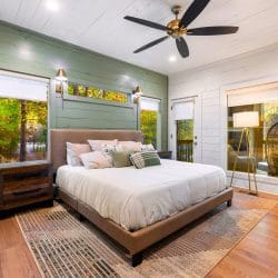King bed on a leather headboard against a sage-green plank wall, with a ceiling fan and windows on both sides