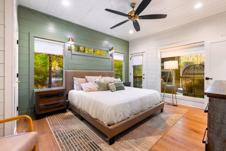 King bed on a leather headboard against a sage-green plank wall, with a ceiling fan and windows on both sides
