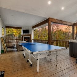 Covered back deck with a ping pong table, a hot tub at the right and a wicker egg chair and outdoor fireplace beyond