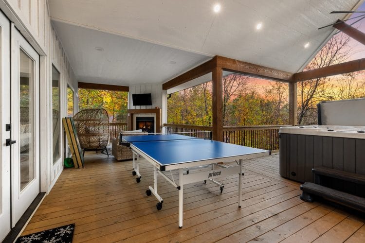 Covered back deck with a ping pong table, a hot tub at the right and a wicker egg chair and outdoor fireplace beyond