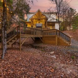 Back of the cabin with a wide wooden deck, stairs and a lit glass peak at dusk