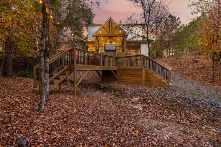 Back of the cabin with a wide wooden deck, stairs and a lit glass peak at dusk