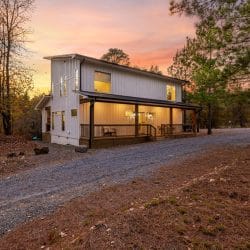Front of the white two-story cabin with a metal roof and long porch, gravel drive curving past at sunset